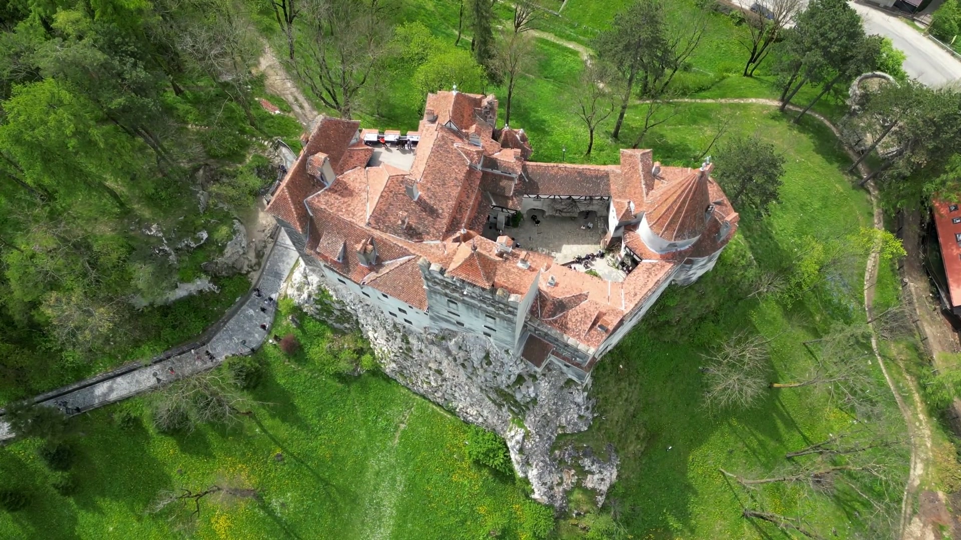 Vedere aeriană oblică foarte înaltă: conturul acoperișurilor, hornuri, curte și stâncă. Cadru real extras la 00:12 din filmul sursă, redus la 1920 × 1080 px, fără sinteză IA.