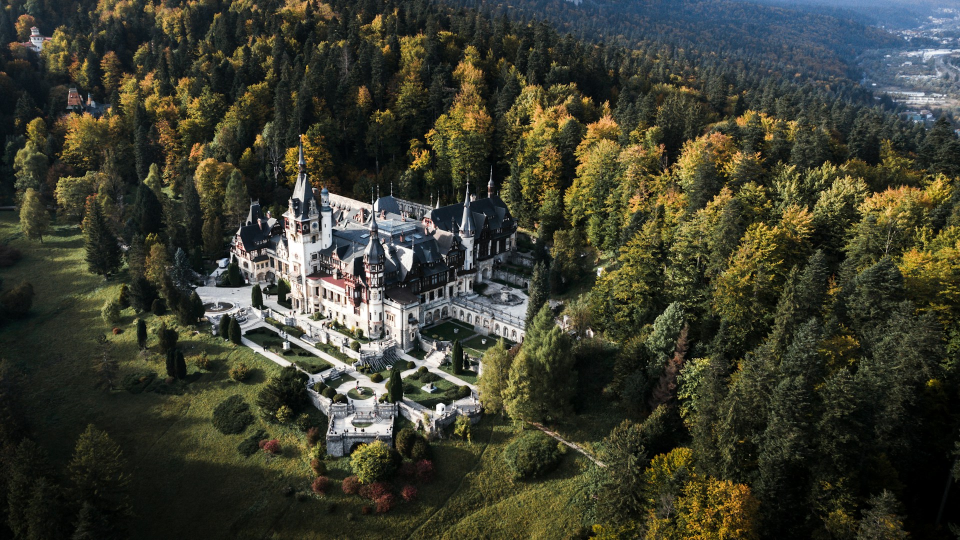 Vedere aeriană dinspre laturile opuse peluzei principale: aripi, turnuri și acoperișuri între arbori.