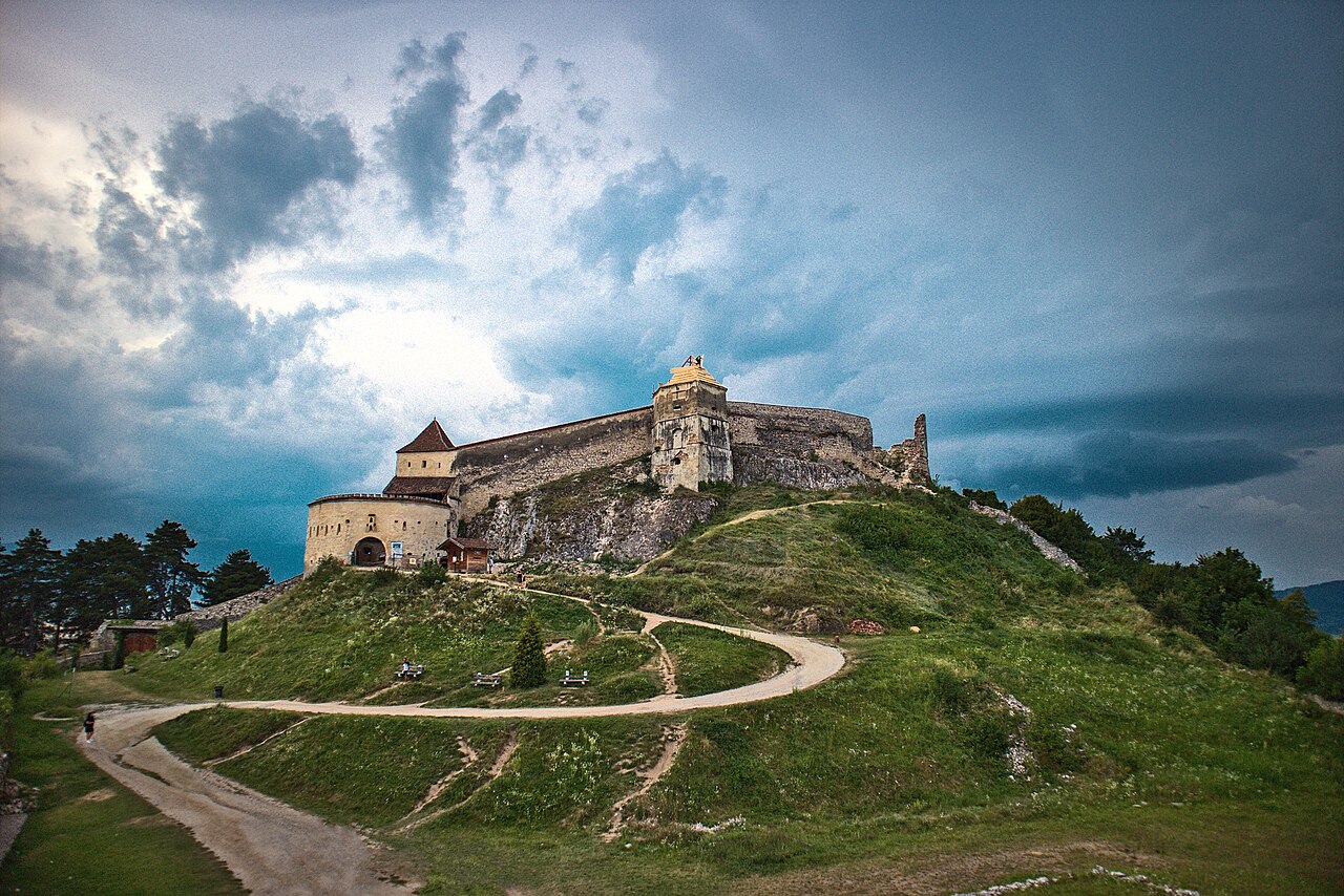 Vedere exterioară largă din zona de acces: ziduri, turnul de poartă și versant.