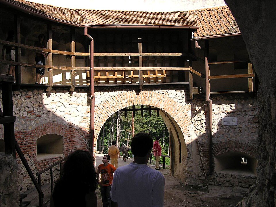 Poarta arcuită din incintă, galeria de lemn și structura acoperișului văzute din curte.