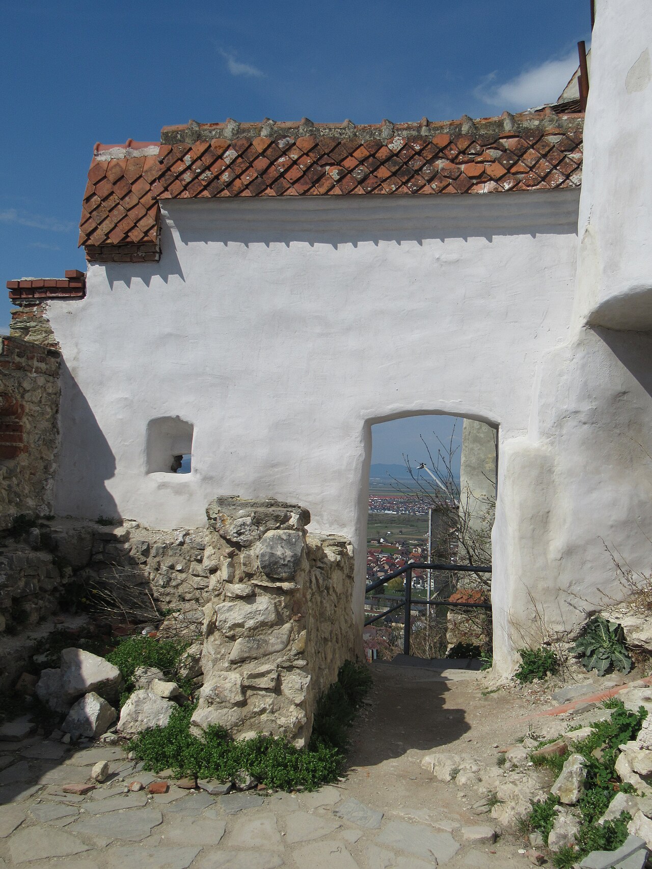 Clădire tencuită alb cu ușă și ferestre, integrată între ruine de piatră; stare diferită de restaurare.