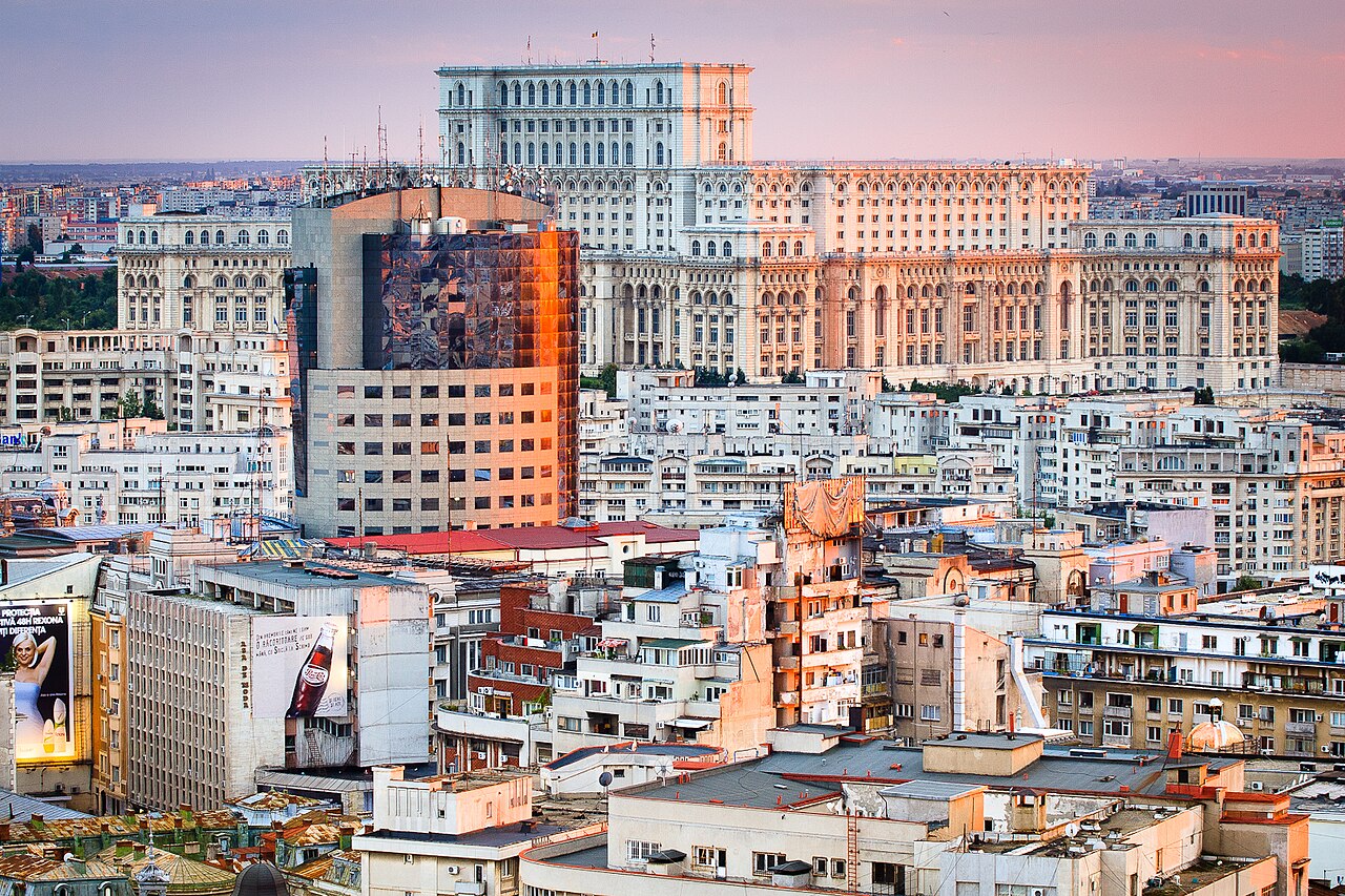 Volumul superior în trepte și ritmul fațadei văzute din oraș; clădiri urbane în prim-plan.