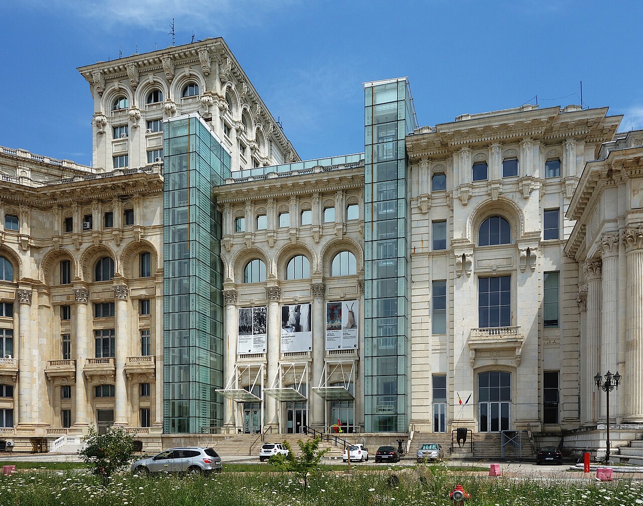 Fațada din zona MNAC, cu liftul exterior de sticlă, turnuri de colț și goluri arcuite.