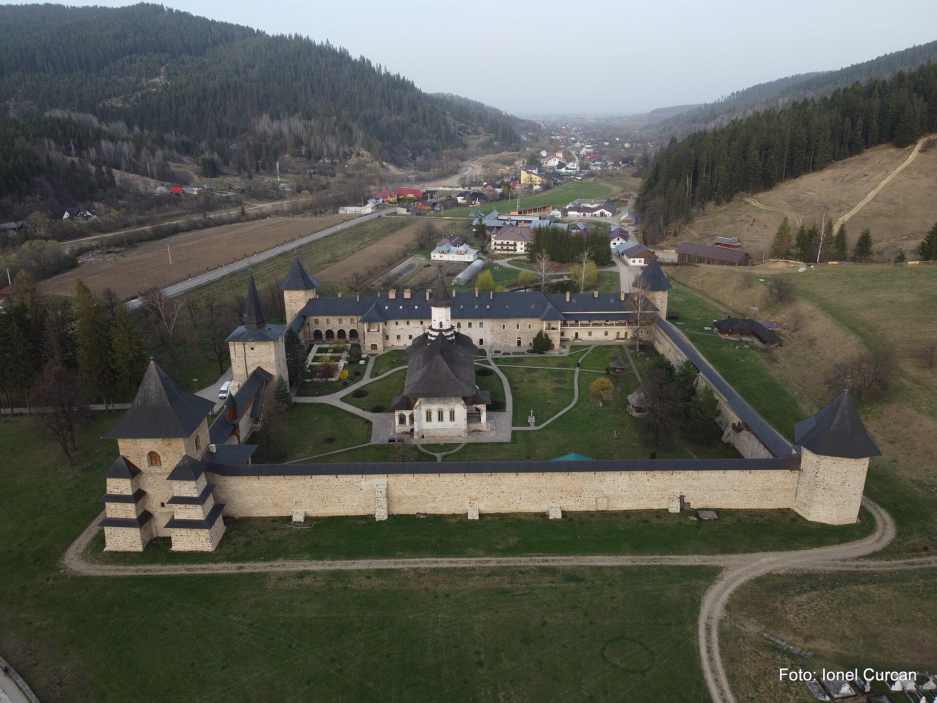 Vedere aeriană oblică din fața unui zid lung: turnuri, biserică și clădiri perimetrale