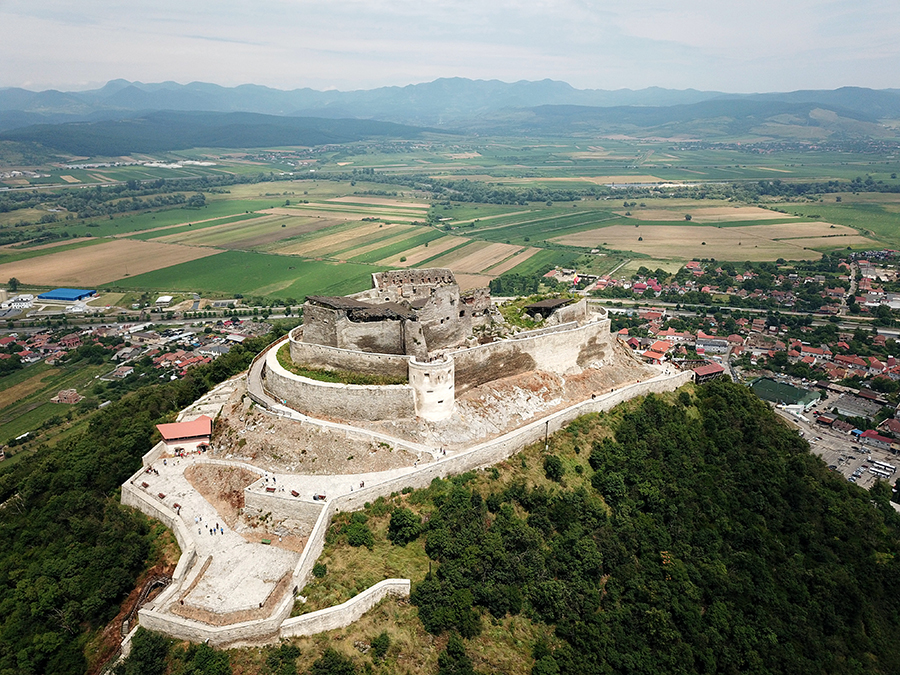 Vedere aeriană oblică a cetății: incinte suprapuse, ruine și dealul din jur