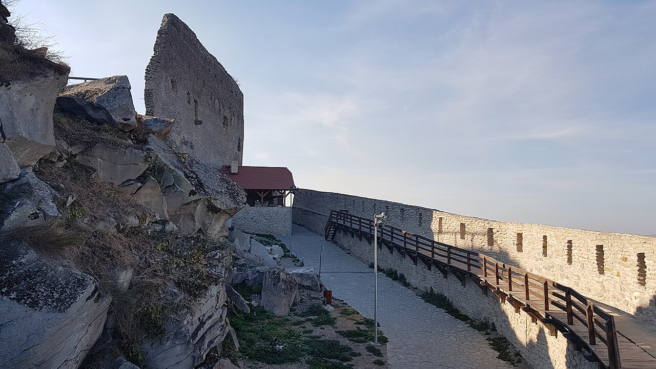 Pasarelă de acces pe lângă ruină și zidul lung cu goluri