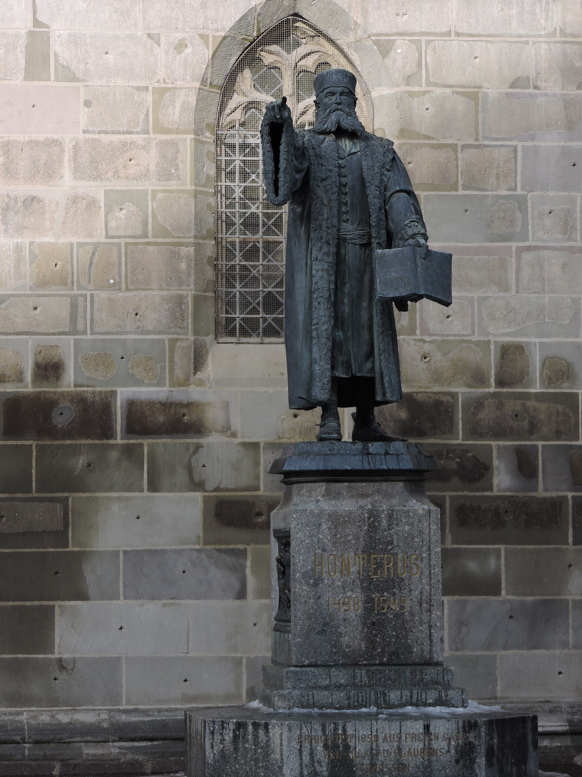 Monumentul lui Johannes Honterus în relație cu zidul și fereastra bisericii.