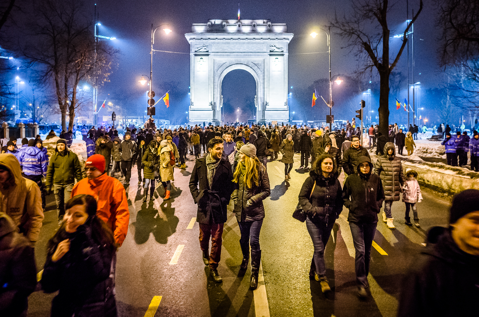 Arcul actual văzut frontal în iluminare nocturnă, cu oameni în prim-plan; silueta și arcada sunt lizibile.