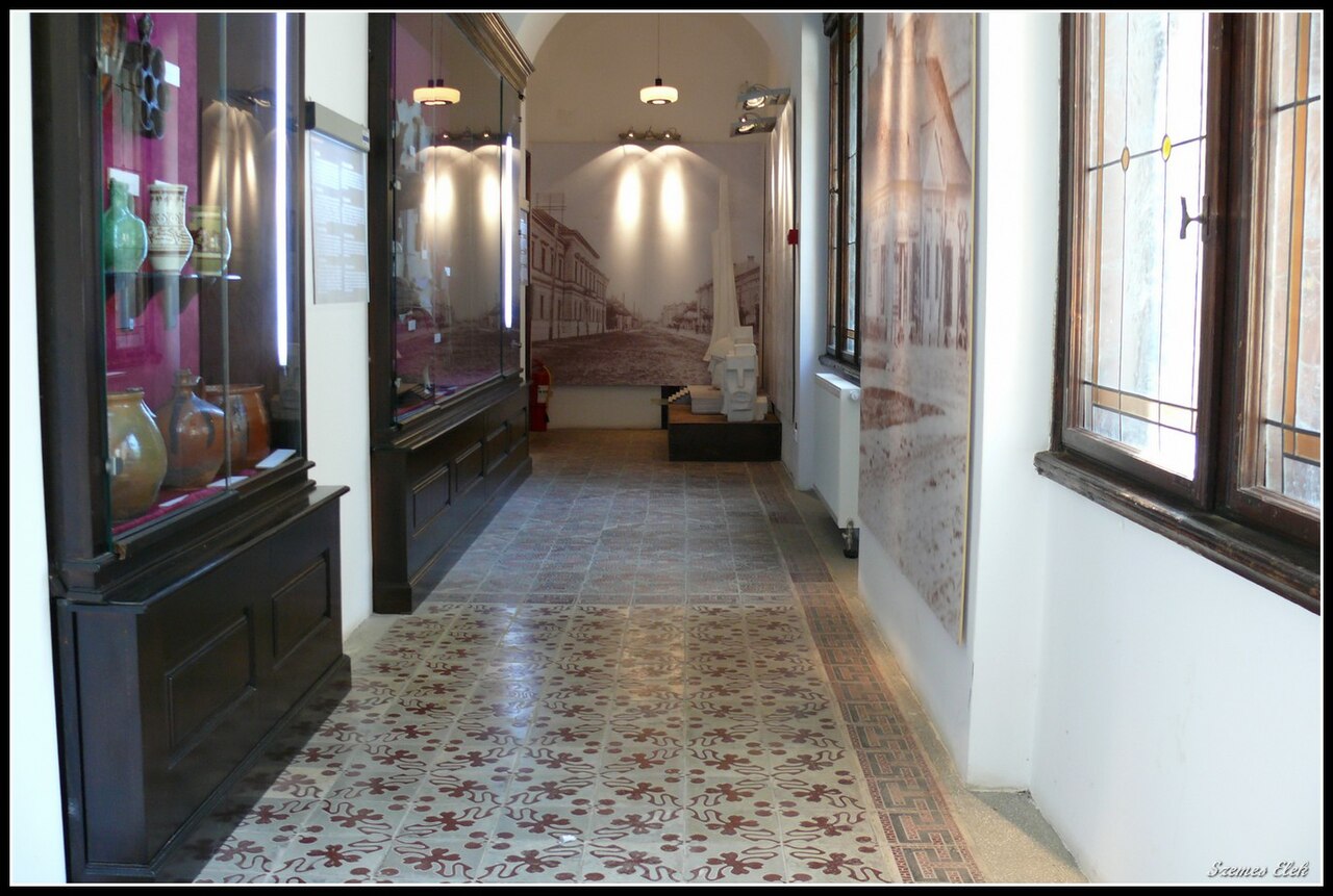 Coridor muzeal cu ferestre, goluri de trecere și pardoseală geometrică.