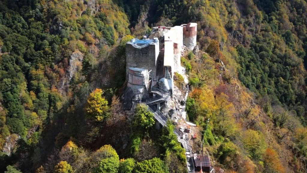 Vedere aeriană oblică dinspre acces: donjon, scări, acoperiri de protecție și creasta stâncoasă.