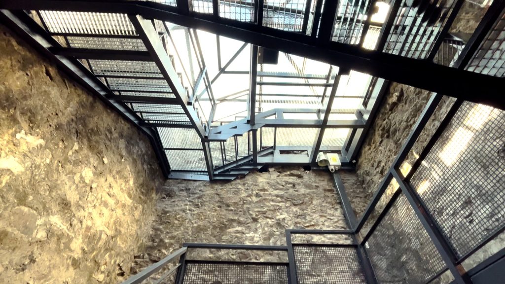 Interiorul unei structuri protejate: ziduri de piatră, scară și acoperire metalică vitrată.