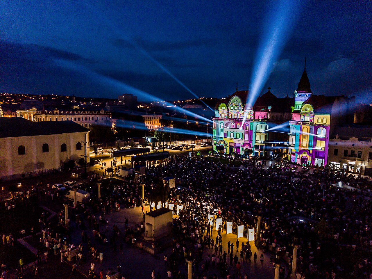 Vedere oblică de la înălțime asupra palatului și pieței în timpul unei proiecții luminoase; acoperișurile sunt în umbră.
