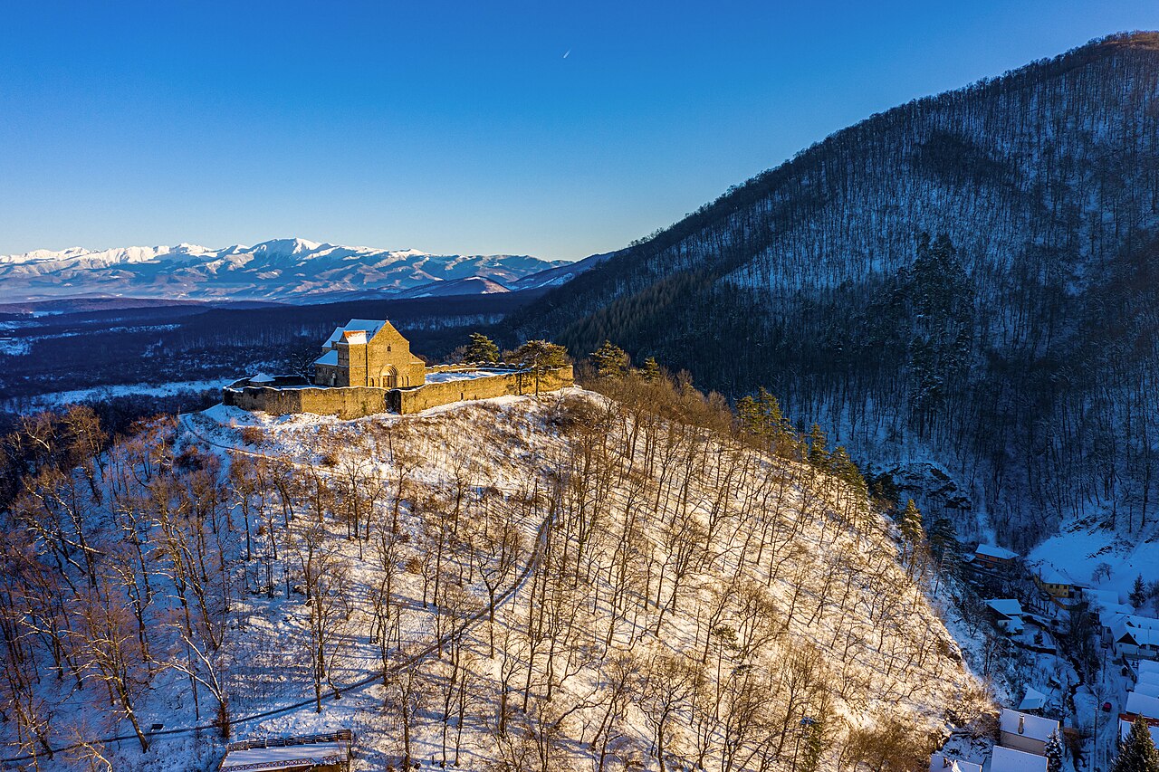Fotografie aeriană dinspre versant, cu zidul circular și biserica pe culme.