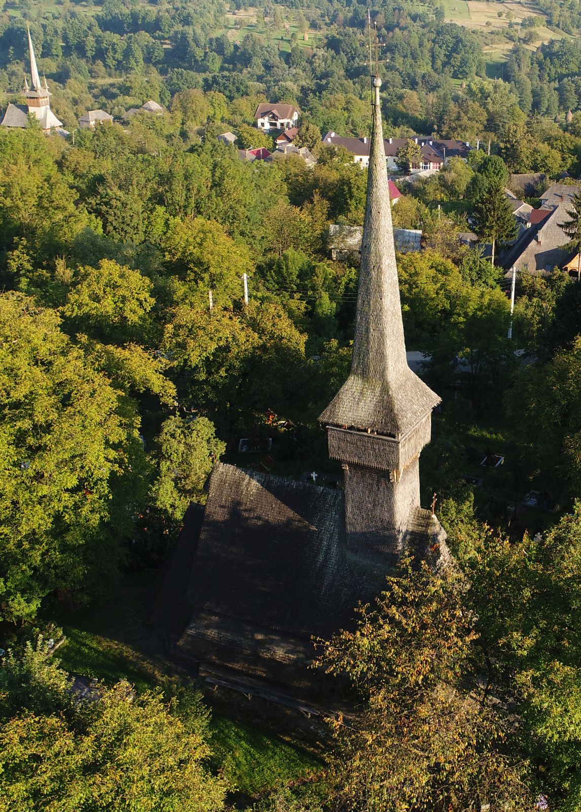 Vedere aeriană oblică a bisericii între arbori, cu turnul și cimitirul.